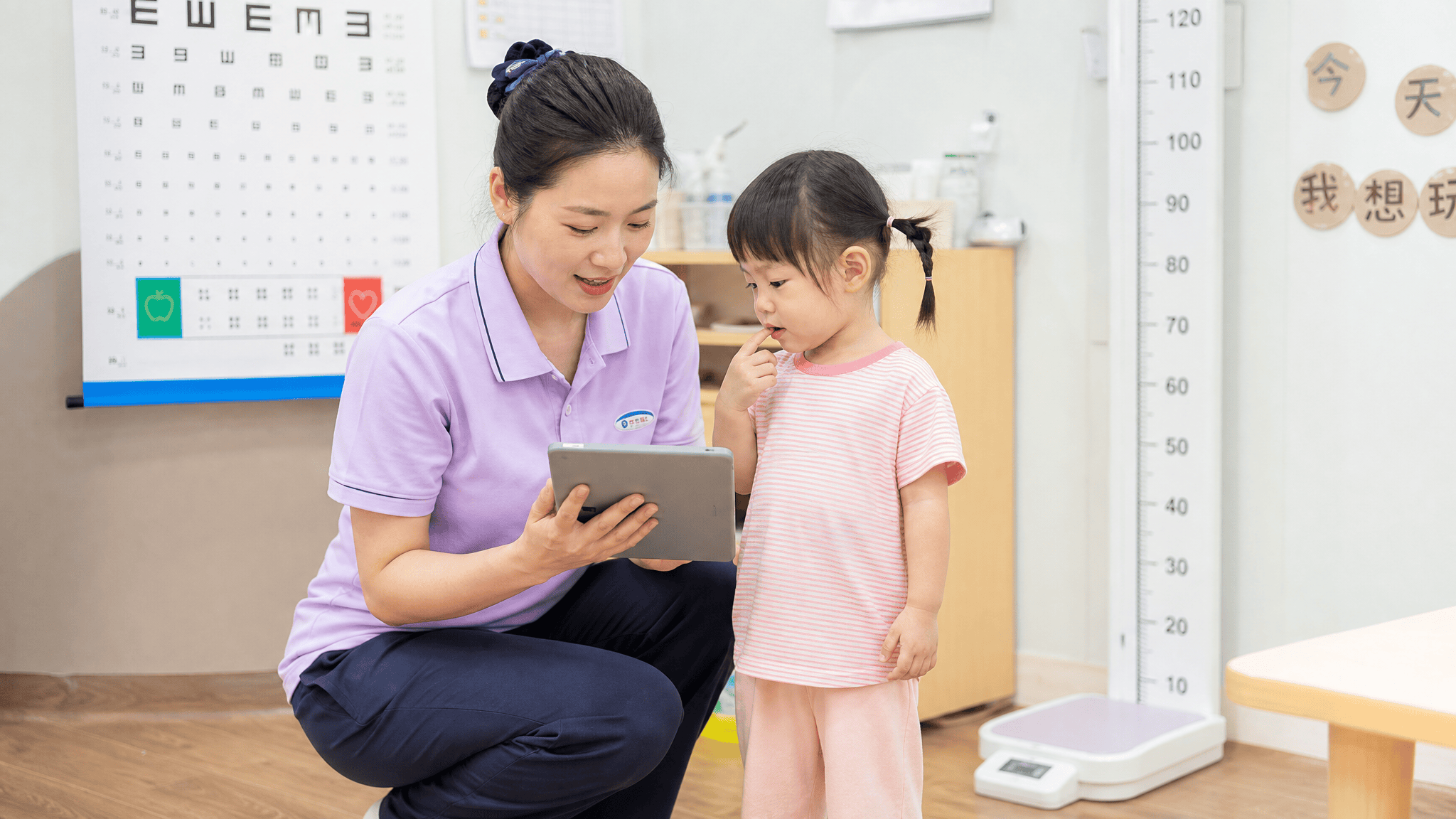托育园所内，保育员蹲在幼儿身边用电子设备记录健康数据，旁边有身高体重测量仪和视力筛查表，明亮温馨的室内环境