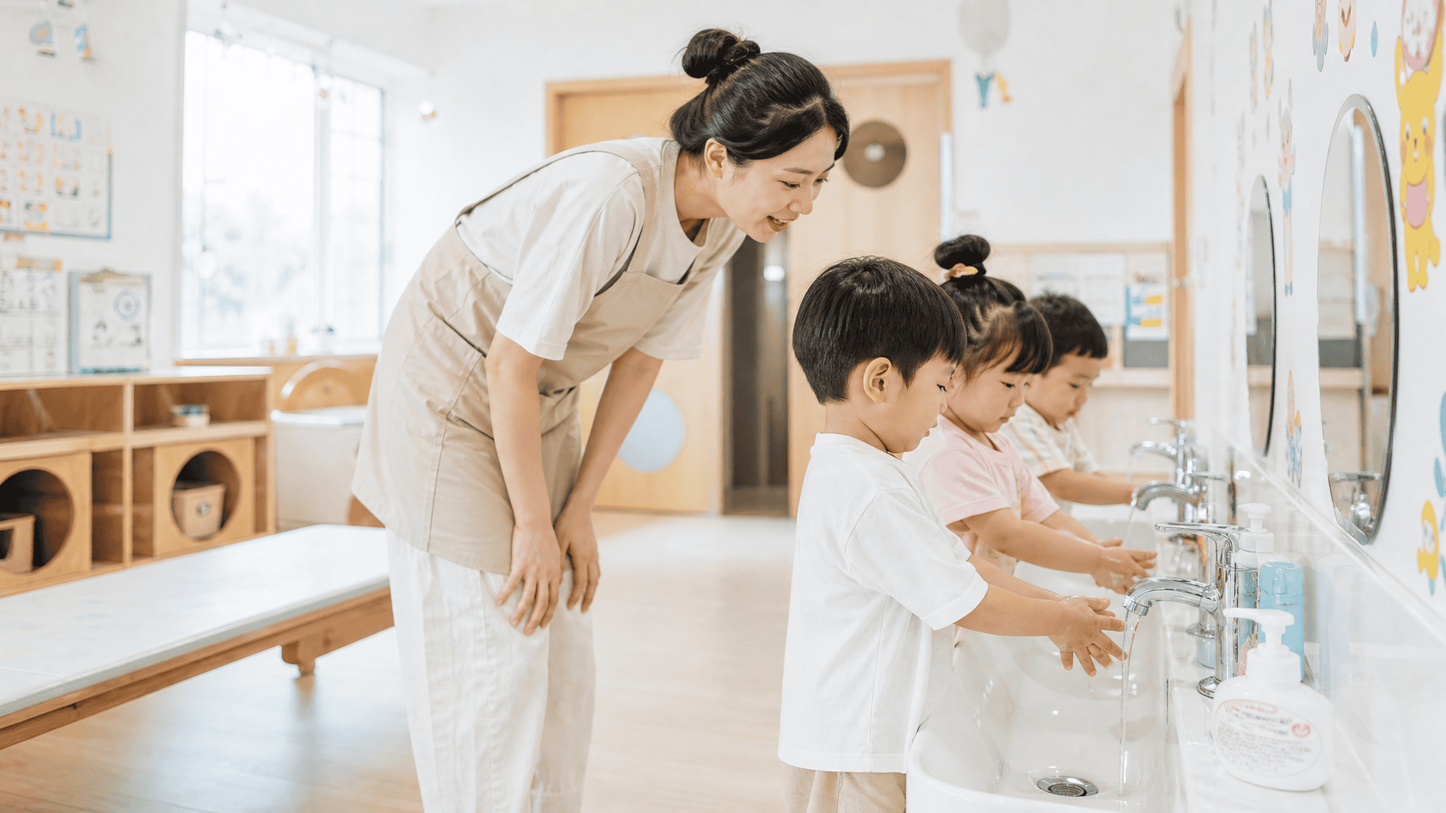 明亮的托育机构盥洗区，几个幼儿在水池前洗手，一位保育员弯腰在旁协助，墙面有卡通贴纸，浅色木质家具